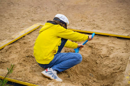 Boy in sandbox diligently digging with blue spade, dressed in jeans, sneakers, yellow hoodie, white cap and trainers.の写真素材