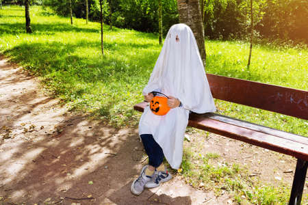 Child in halloween white ghost costume in jeans and sneakers with a basket of pumpkin sitting on park benchの写真素材
