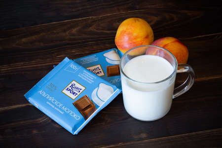 Ritter Sport milk chocolate, two chocolate bars in blue package, glass mug with milk, two noctarines. Still life on a dark wooden surface. - Saint Petersburg, Russia, August 2020のeditorial素材