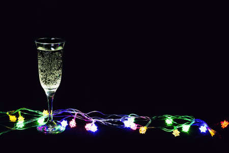 Wineglass of bubbly champagne and glowing multicolored garland on black background.