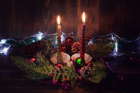 Two burning Christmas candles in wreath of fir branches with cones, luminous garland on dark wooden backgroundの写真素材