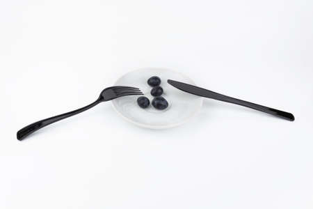 Black fork and knife lying on white porcelain saucer with grapes on white table, plant-based diet.