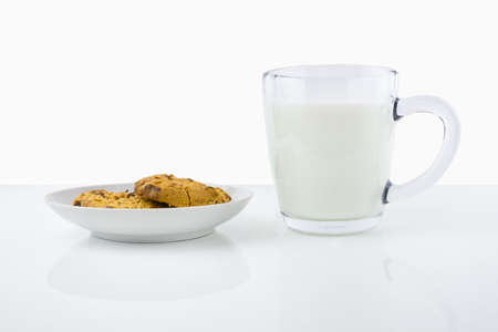 Two American cookies on white porcelain saucer and transparent cup of milk on white surfaceの写真素材