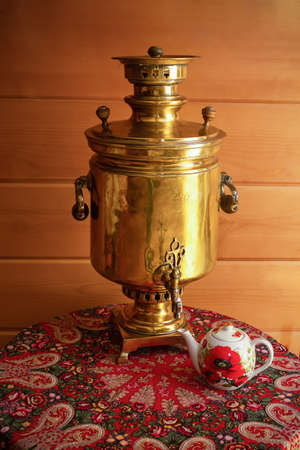 Yellow traditional Russian samovar on table with variegated red tablecloth and teapot. Against background of wooden plank wall.の写真素材