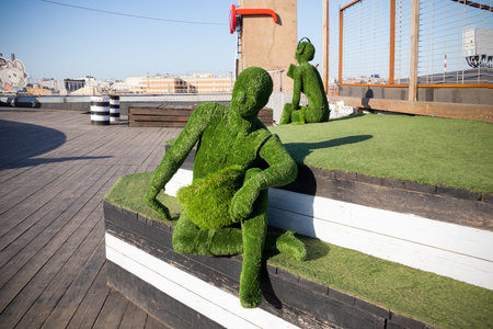 Male figure covered with artificial green grass sitting on steps and female figure with headphones in the background. Outdoor. - Loft project Floors Etagi, Saint Petersburg, Russia, April 2021のeditorial素材