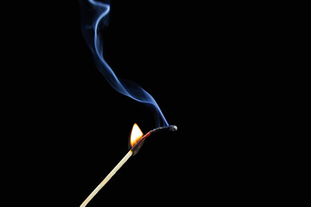 Burning match half burnt with blue plume of smoke on black background.