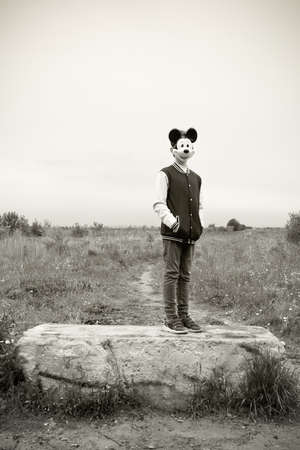 Teenage in Mickey Mouse mask, college jacket and jeans standing on stone in field, toned in sepia....