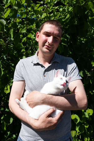 Man in gray T-shirt holding a white cat in his hands on background of green leaves illuminated by sunlight.の写真素材