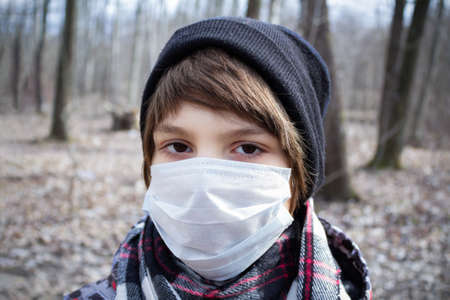 Portrait of boy in disposable medical mask, scarf and hat against a background of trees.の写真素材