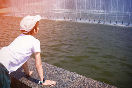 Boy leaning on the fountain side and looking at the water, wearing white T-shirt and cap, view from the backの写真素材