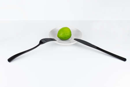 Fresh lime on white plate with black fork and knife on white surface