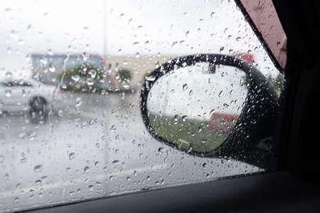 Car glass covered with raindrops and side mirror, view of rainy street through car window