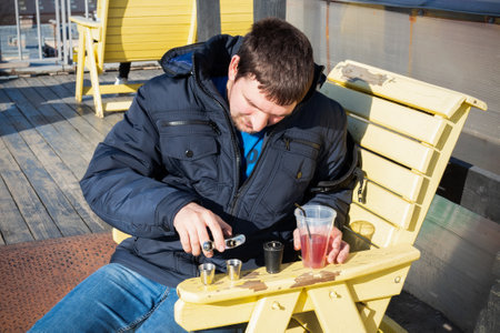 Young man pouring alcohol from flask, sitting on wooden yellow chair on sunny terrace in blue jeans...