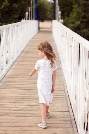 back view. A little girl in a white dress with flowing hair walks along a wooden bridge.の写真素材