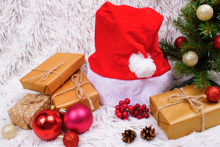 Many presents wrapped in a craft paper, Santa hat and decorative accessories under the decorated Christmas tree on white fur background.の写真素材