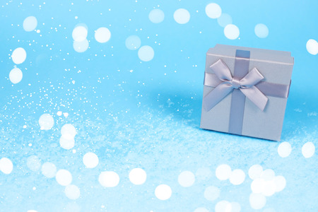Beautiful gift box of silver color with cute little bow made of silky ribbon on blue background decorated with festive lights and snow with copy space. Holiday concept.の写真素材
