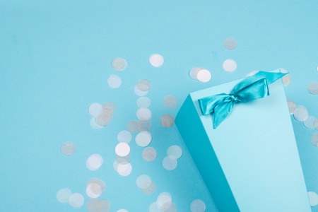 Blue box with shiny satin ribbon tied in a bow with scattered pastel confetti on the background of the same tone. Trendy creative celebration backdrop with copy space for text.の写真素材