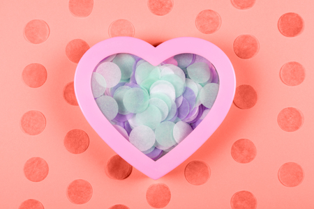 Tender heart-shaped box full of colorful round paper confetti on decorated coral background. Valentine's Day and romantic concept. Love symbols. Festive backdrop with copy space.の写真素材