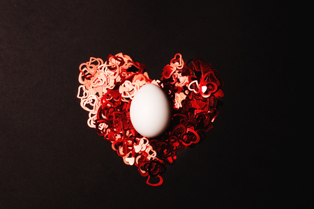One white egg on a heart of vibrant red heart-shaped confetti on black background. Selective focus. Easter and holiday concept.の写真素材