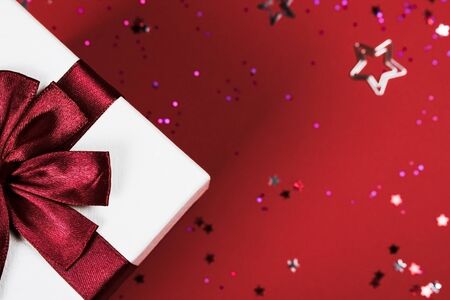 Close-up of white gift box with siljy shiny ribbon on red background decorated with glitter confetti...
