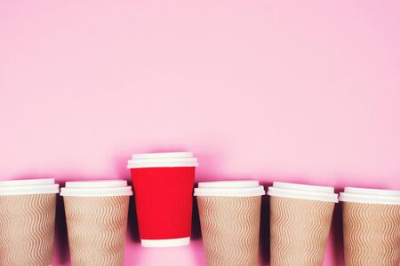Many brown and one red paper coffee cups with white plastic covers laid in a row on pink background...