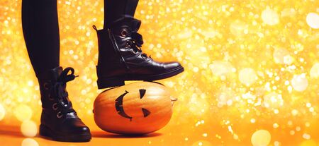 Witch legs of girl in black pants and leather shoes standing a scary pumpkin with spooky face on orange background for Halloween concept, decorated with festive magic lights with copy space for text.の写真素材