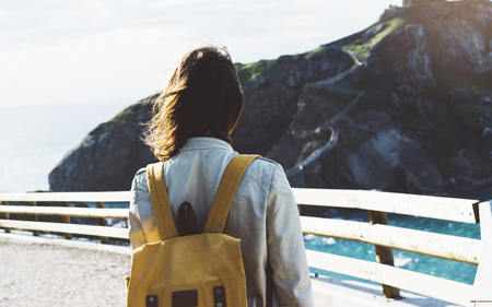 Hipster young girl with backpack enjoying sunset on seascape on peak mountain. Tourist traveler on background valley landscape view mockup. Hiker looking sunlight ocean in trip holiday in basque islandの写真素材