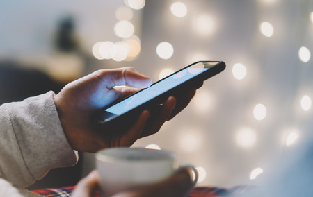 Hipster girl using mobile phone and drink coffee in home atmosphere, person holding smartphone on background glow bokeh Christmas illimination, female hands texting on relax glitter xmas decoration, mockup blurの写真素材