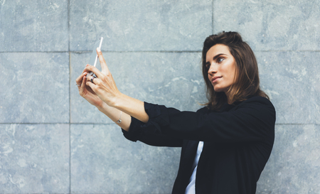 Portrait young businesswomen in black suit using smartphone isolated on background concrete gray wall mockup, pretty hipster manager holding mobile gadget,  girl making selfie photo, connectの写真素材