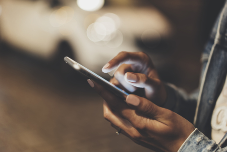 Girl pointing finger on screen smartphone on background auto taxi, illumination bokeh color light in night atmospheric city, hipster using in female hands and texting mobile phone, mockup glitter street lifestyleの写真素材