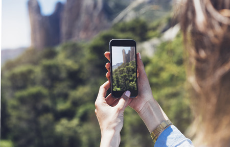 Hipster girl making photo on smartphone mobile closeup, view tourist hands using gadget phone in travel on background mountains and sky landscape; finger touch screen cellphone mockup nature, templatesの写真素材