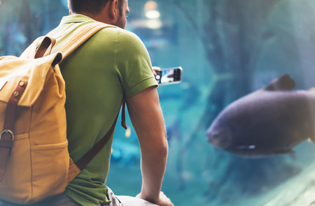 Hipster with yellow backpack, map, phone. View from back tourist traveler on background blue sea aquarium. Person hiker in oceanarium museum Barcelona and photographer pictures fish smartphone mockupの写真素材