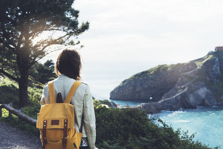 Hipster young girl with backpack enjoying sunset on seascape on peak mountain. Tourist traveler on background valley landscape view mockup. Hiker looking sunlight ocean in trip holiday in basque islandの写真素材