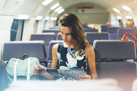Enjoying travel. Young pretty woman traveling by train sitting near the window using smartphone and looking map. Tourist texting online message and plan route of railway, railroad transport conceptの写真素材