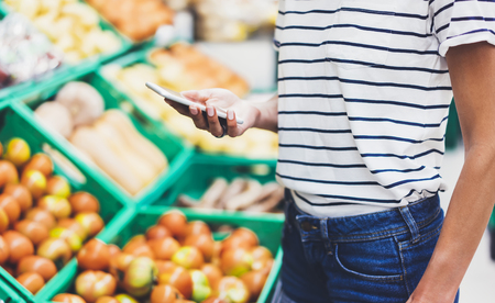 Young woman shopping healthy food in supermarket blur background. Female hands buy products tomato using smartphone in store. Hipster at grocery using smartphone. Person comparing price of produceの写真素材
