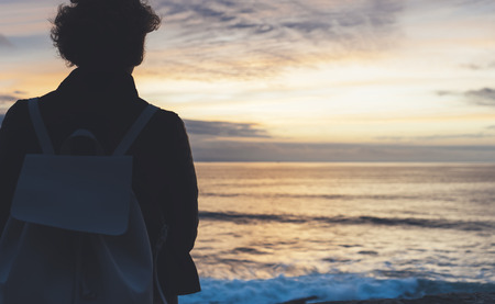 Young hipster relax on the beach on background ocean sunrise, silhouette romantic person looking view evening seascape, girl enjoy sunset on coast, travel holidays vacation, back figure on backdrop seaの写真素材