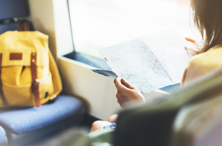 Enjoying travel. Young pretty woman traveling by train sitting near the window holding and looking map. Tourist plan route of railway, railroad transport conceptの写真素材