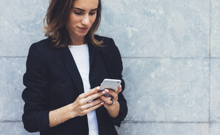 Portrait young businesswomen in black suit using smartphone isolated on background concrete gray wall mockup, pretty hipster manager holding mobile gadget, girl smile texting message, connectの写真素材