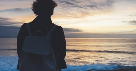 Young hipster relax on the beach on background ocean sunrise, silhouette romantic person looking view evening seascape, girl enjoy sunset on coast, travel holidays vacation, back figure on backdrop seaの写真素材
