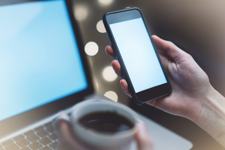 Hipster girl using computer and cup coffee in home atmosphere, person holding blank smartphone on background glow illumination, female hands texting on relax glitter bokeh, mockup templates, blur の写真素材