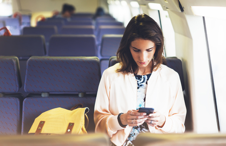 Enjoying travel. Young pretty woman traveling by train sitting near the window using smartphone and looking map. Tourist texting online message and plan route of railway, railroad transport conceptの写真素材