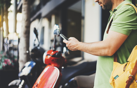 Man pointing on screen smartphone on background red retro scooter, tourist hipster waiting and using in hands mobile phone, traveler connect wifi internet, backdrop summer street in city, mockupの写真素材