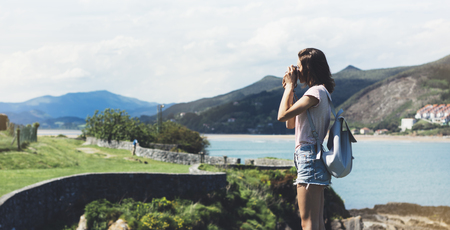 Tourist traveler photographer making pictures sea scape on vintage photo camera on background ocean, hipster girl enjoying green peak mountain and nature holiday, mock up waves view, blurred backdropの写真素材
