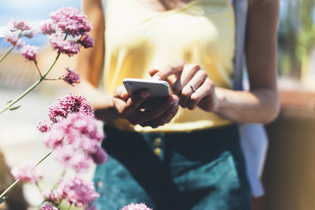 Hipster text message on smart phone or technology, mock up of blank screen. Young girl using cellphone on color flower background close. Tourist female hands holding gadget on blurred summer backdropの写真素材