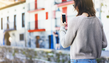 Girl traveler making photo screen smart phone on background old color house in summer sun atmospheric city, young hipster tourist using in female hands mobile phone, mock up flare street, content lifestyleの写真素材