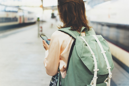 Enjoying travel. Young pretty woman waiting on the station platform with backpack on background electric train using smartphone. Tourist texting message and plan route of railway, railroad transport conceptの写真素材
