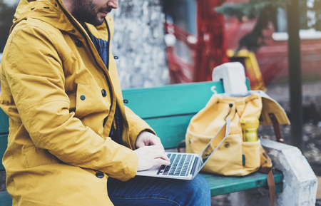 Hipster with yellow backpack, jacket, cap, thermo cup of coffee using computer open laptop in spring street outdoor, tourist man typing on keyboard, traveler connect wifi internet, freelance process in workspaceの写真素材