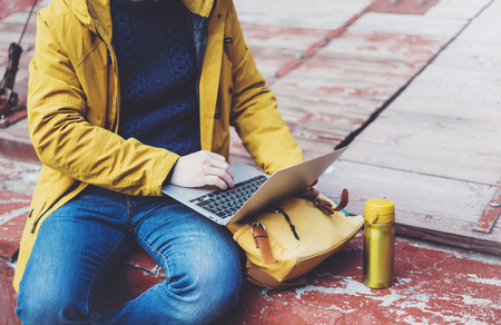 Hipster with yellow backpack, jacket, cap, thermo cup of coffee using computer open laptop in spring street outdoor, tourist man typing on keyboard, traveler connect wifi internet, freelance process in workspaceの写真素材