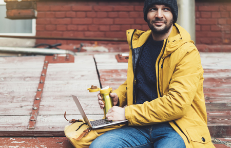 Hipster with yellow backpack, jacket, cap, drink coffee of thermo cup, freelance using computer open laptop in spring street outdoor, tourist man typing on keyboard, traveler connect wifi internet, process in workspaceの写真素材