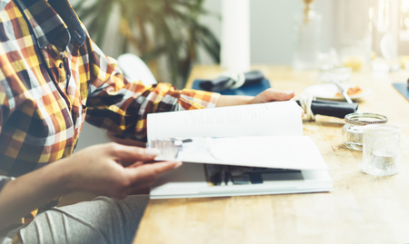 Young girl reads book during breakfast and coffee, female hands close up flipping through magazine pages in home relax atmosphere room on background of natural wooden table, woman is studying, mockの写真素材